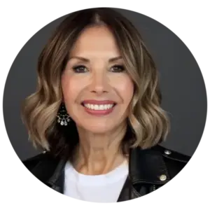 Smiling woman with shoulder-length wavy light brown hair, wearing a black leather jacket and white top, against a dark gray background.